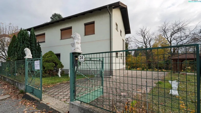 Einfamilienhaus mit heller Fassade, grüner Zaunanlage und gepflasterter Auffahrt. Der Gartenbereich umfasst Bäume und einen kleinen Pavillon.