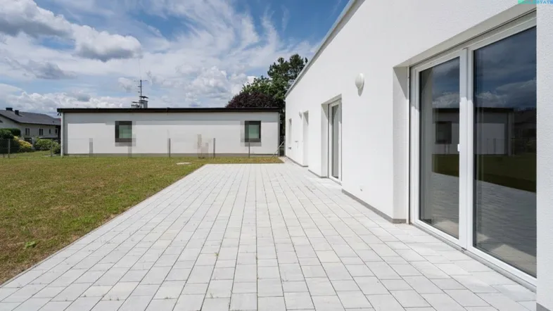 Moderne weiße Fassade mit großer gepflasterter Terrasse und angrenzendem Rasenbereich unter blauem Himmel.