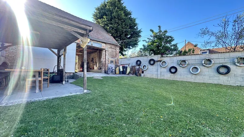 Sonniger Garten mit Rasenfläche, überdachtem Sitzplatz und Blick auf ein Backsteingebäude.