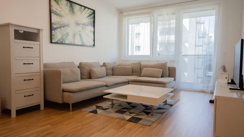 Helles Wohnzimmer mit beigem Ecksofa, Holzboden und Zugang zu einem Balkon.