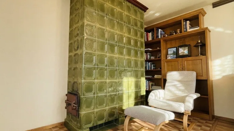 Gemütliches Wohnzimmer mit traditionellem grünem Kachelofen, Lesesessel und Holzregal.