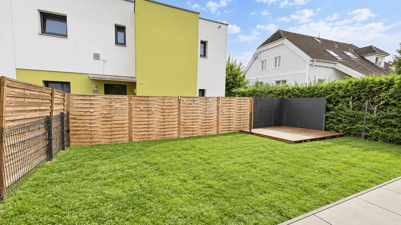 Gepflegter Gartenbereich mit Rasenfläche, Holzzaun und einer kleinen Holzterrasse hinter dem Haus.