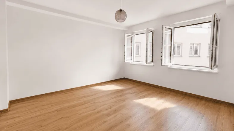 Helles, leeres Zimmer mit Holzboden und geöffneten Fenstern, die auf ein Gebäude blicken.