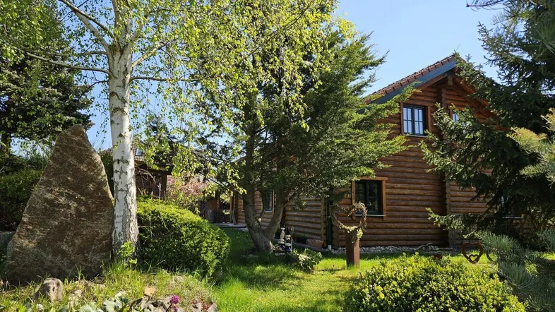 Ein charmantes Blockhaus umgeben von einem gepflegten Garten mit Bäumen und einem großen dekorativen Stein an einem sonnigen Tag.