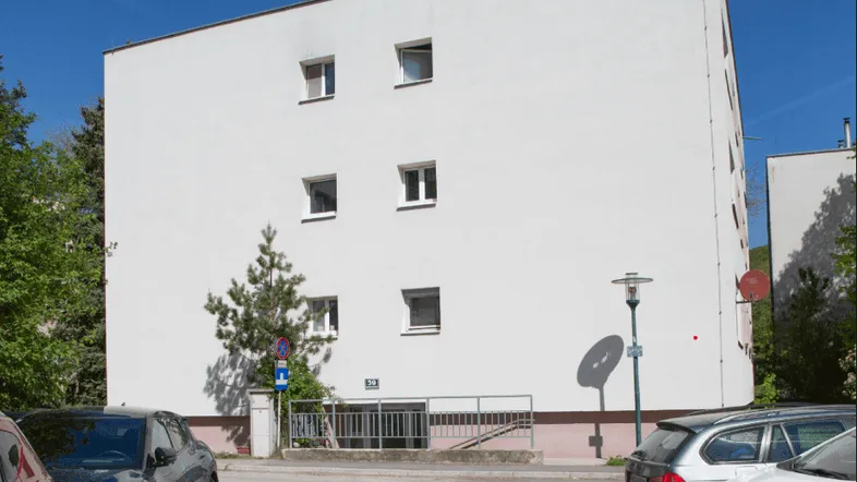 Weißes Mehrfamilienhaus mit schlichter Fassade und Parkplätzen im Vordergrund unter blauem Himmel.