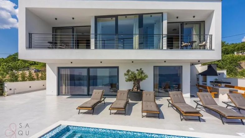 Moderne Villa mit Pool, Sonnenliegen und Balkonen, umgeben von grüner Landschaft unter blauem Himmel.