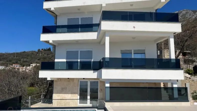 Moderne weiße Villa mit Glasbalkonen und einem Pool vor einer Bergkulisse.