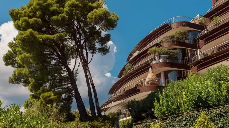 Elegantes Gebäude mit geschwungenen Balkonen, umgeben von üppiger mediterraner Vegetation und einem Weg zum Meer.