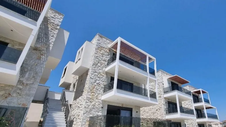 Moderne Architektur mit Steinfassade, mehreren Balkonen und Holzpergolen unter blauem Himmel.