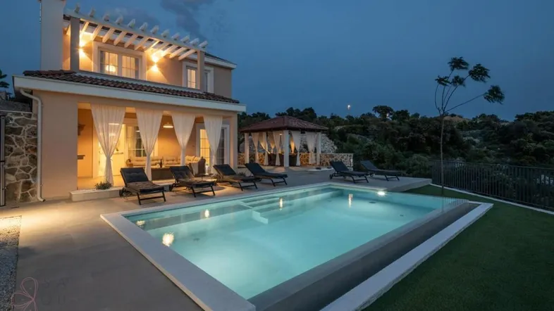 Elegante Villa bei Nacht mit beleuchtetem Poolbereich, umgeben von üppiger Vegetation und einem Pavillon.