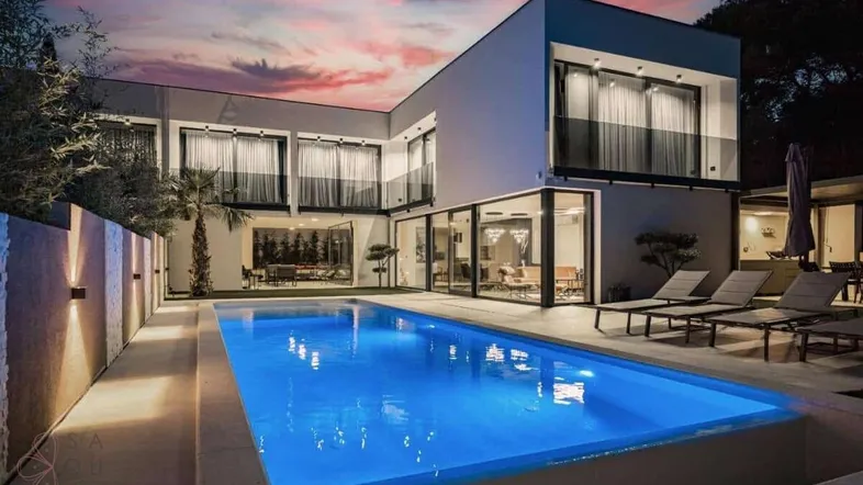 Moderne Villa mit beleuchtetem Pool und großen Glasfronten bei Sonnenuntergang.