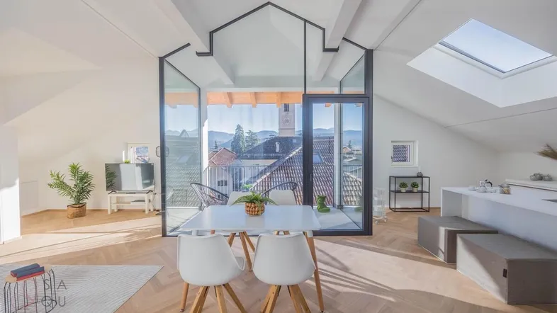 Helles Dachgeschoss-Apartment mit offenem Wohn-Essbereich, Holzbalken und Zugang zum Balkon mit Stadtblick.