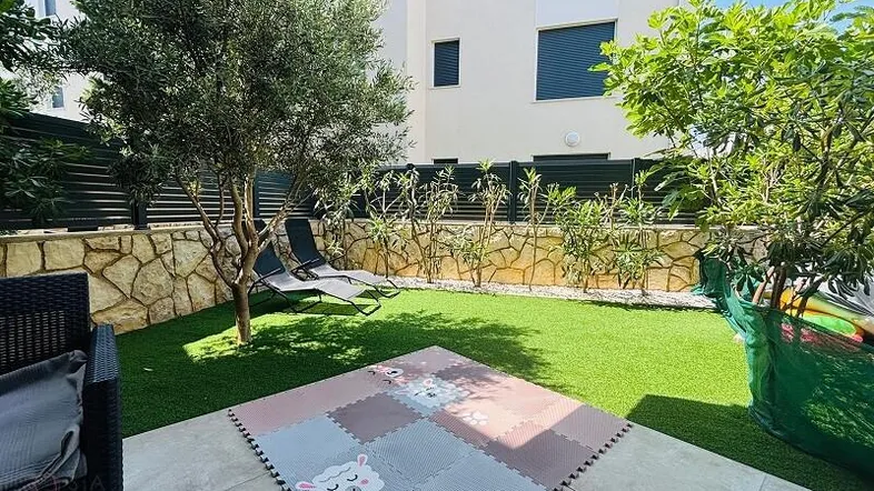 Gepflegter Gartenbereich mit Kunstrasen, Olivenbaum und Liegestühlen, ideal zum Entspannen und Spielen.
