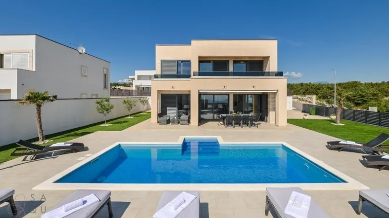 Moderne Villa mit großem Swimmingpool, Sonnenterrasse und Liegestühlen unter blauem Himmel.