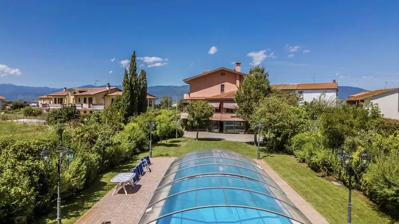 Poolbereich mit überdachtem Schwimmbecken und Liegestühlen in einer grünen Gartenanlage.