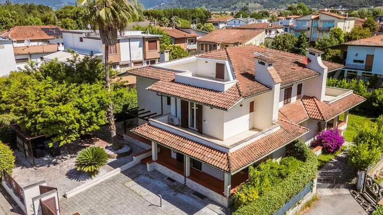 Luftaufnahme eines Einfamilienhauses mit Ziegeldach, umgeben von viel Grün und Bergen im Hintergrund.
