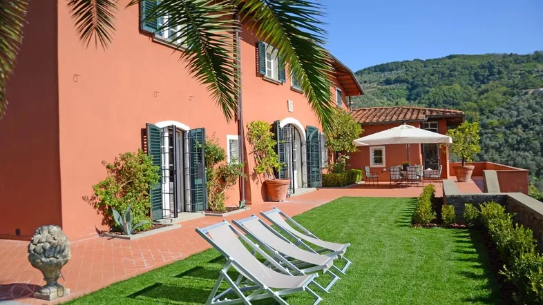 Mediterranes Haus mit ockerfarbener Fassade, gepflegtem Rasen und Sonnenliegen im Gartenbereich.