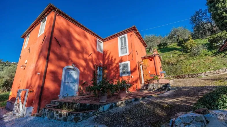 Traditionelles Landhaus mit leuchtend oranger Fassade und blauem Himmel, umgeben von mediterraner Vegetation.