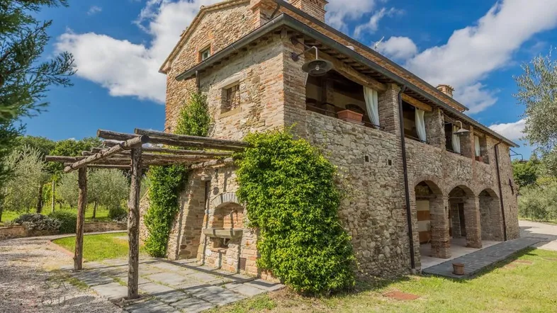 Traditionelles Landhaus aus Stein und Ziegeln mit Pergola und üppigem Grün unter blauem Himmel.