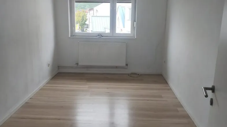 Leerer, heller Raum mit Laminatboden, Fenster mit Rollladen und Heizkörper.