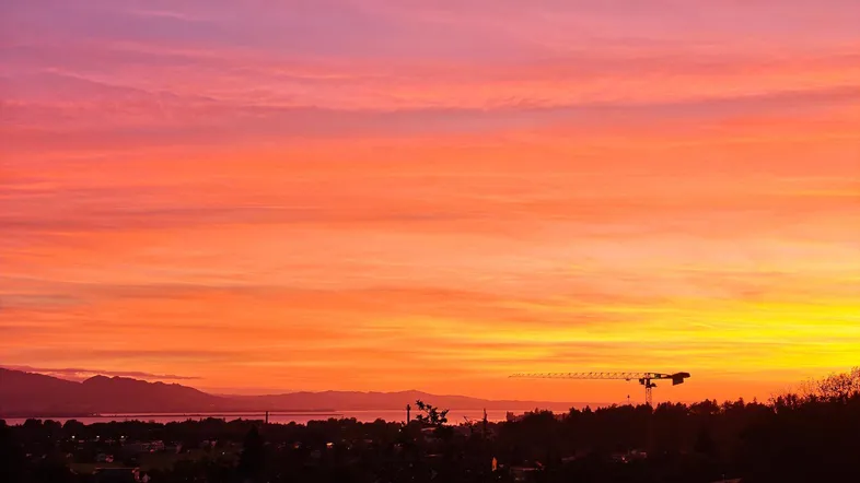 Spektakulärer Sonnenuntergang über der Landschaft mit See, Bergen und einem Kran im Hintergrund.