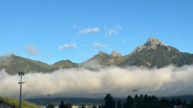 Panoramablick auf majestätische Berge, teilweise von Nebel umhüllt, unter einem klaren blauen Himmel.