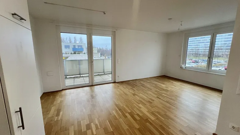 Geräumiger, leerer Wohnbereich mit Parkettboden und Zugang zu einem Balkon.