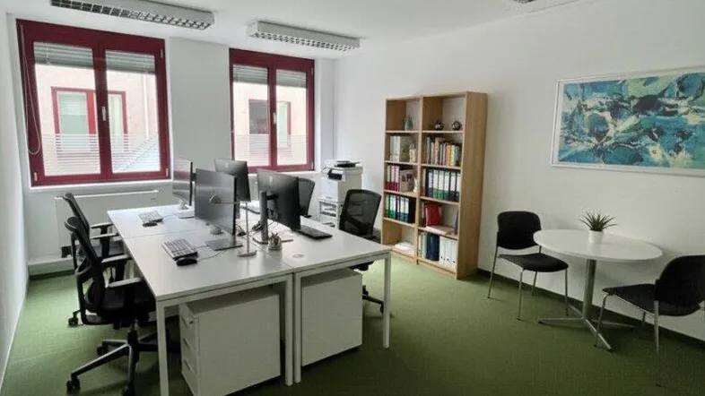 Helles Büro mit grüner Teppichboden, mehreren Arbeitsplätzen und Fenstern mit rotem Rahmen.