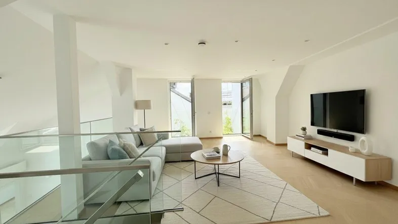Helles Wohnzimmer mit modernem Sofa, Fernseher und Zugang zu einem Außenbereich, gekennzeichnet durch eine Glasbrüstung.