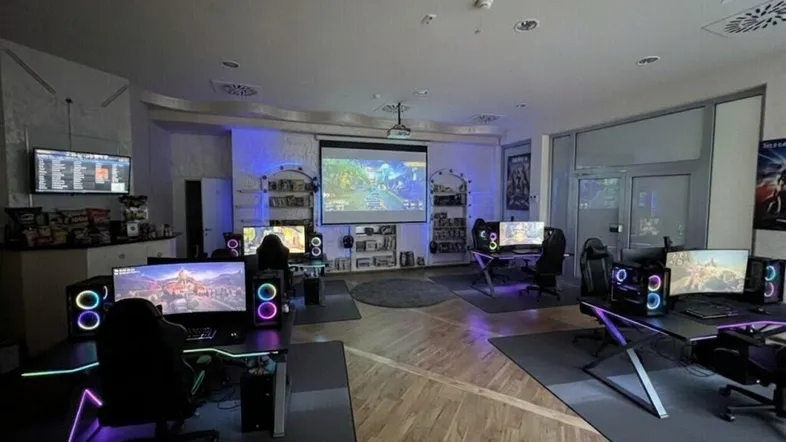 Geräumiger Gaming-Raum mit mehreren modernen Spielstationen, großem Projektorbildschirm und atmosphärischer Beleuchtung.