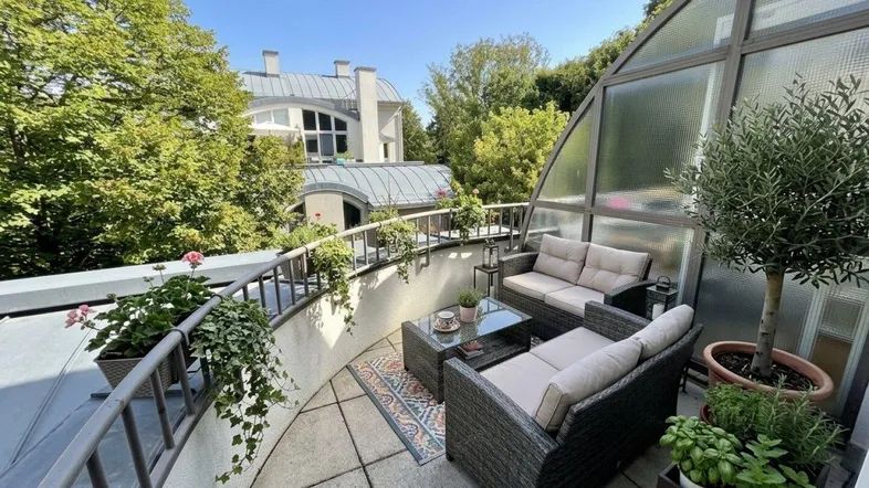 Eine möblierte Terrasse mit Pflanzen und einem verglasten Bereich, umgeben von Bäumen und Gebäuden.