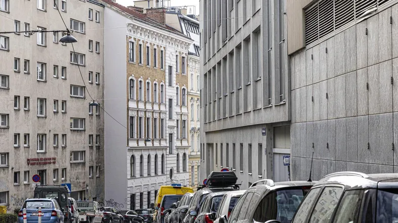 Eine belebte Straße mit parkenden Autos und verschiedenen Gebäudefassaden in einer städtischen Umgebung.