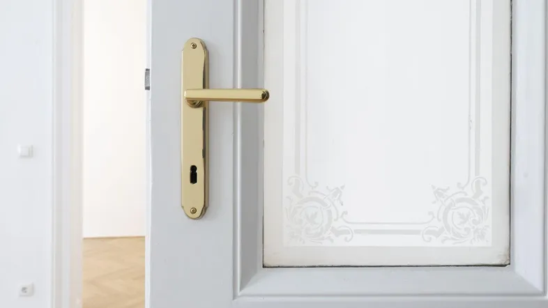 Nahaufnahme einer weißen Tür mit goldener Klinke und dekorativem Milchglas, die in einen hellen Raum führt.
