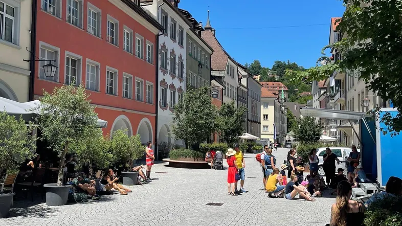 Belebter Stadtplatz mit historischen Gebäuden, Kopfsteinpflaster und Menschen, die die Sonne genießen.