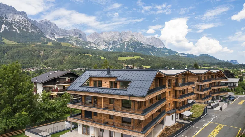 Moderne Holzfassade eines Mehrfamilienhauses mit Balkonen und beeindruckendem Bergblick unter blauem Himmel.