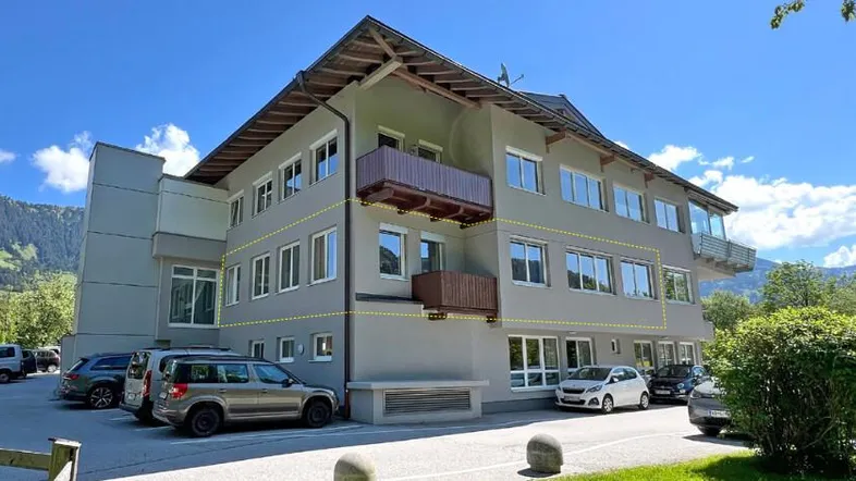 Modernes Mehrfamilienhaus mit mehreren Balkonen und Parkplätzen in bergiger Umgebung.