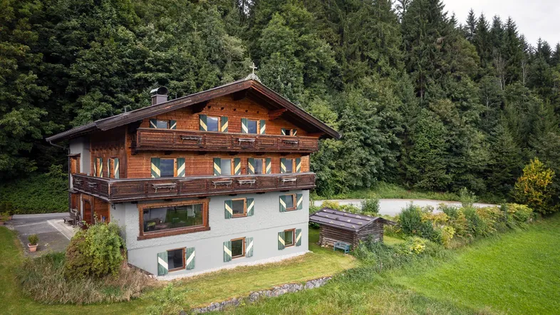 Traditionelles Alpenhaus mit Holzbalkonen und grünen Fensterläden, umgeben von Wald und Wiesen.