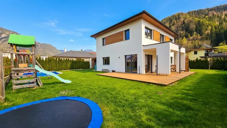 Modernes Einfamilienhaus mit großem Garten, Terrasse und Spielbereich für Kinder vor Bergkulisse.