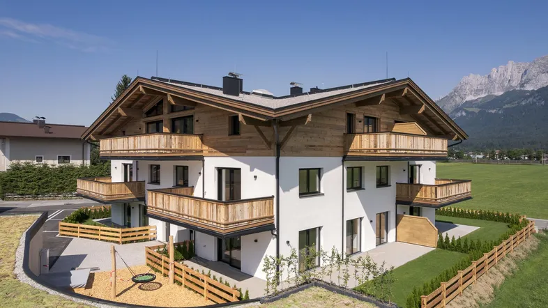 Modernes Mehrfamilienhaus mit Holzfassade, Balkonen und Bergblick, umgeben von Grünflächen und einem kleinen Spielplatz.