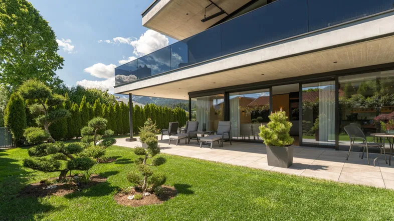 Moderne Villa mit großer Terrasse, Garten und gepflegter Rasenfläche unter blauem Himmel.