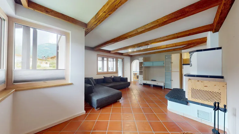 Helles Wohnzimmer mit sichtbaren Holzbalken, Terrakottafliesen und einem traditionellen Kachelofen.