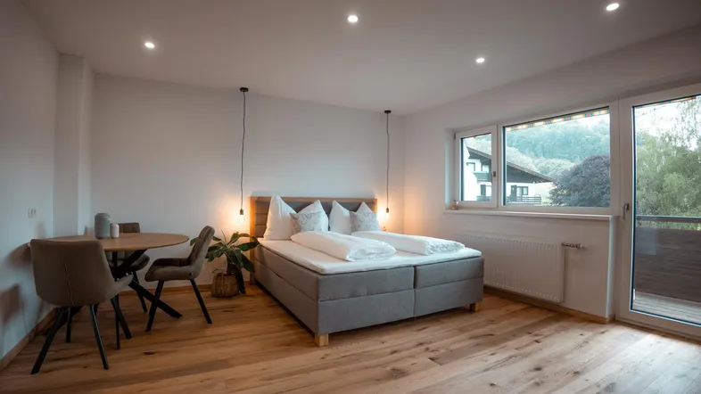 Helles Schlafzimmer mit Holzboden, Doppelbett, Esstisch und Zugang zu einem Balkon.