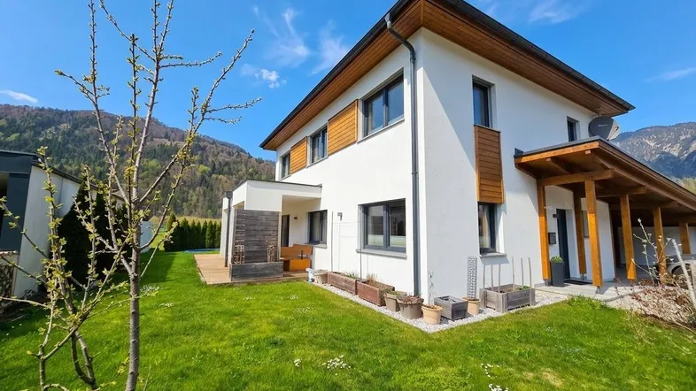 Modernes Einfamilienhaus mit weißer Fassade, Holzdetails und gepflegtem Garten in alpiner Umgebung.