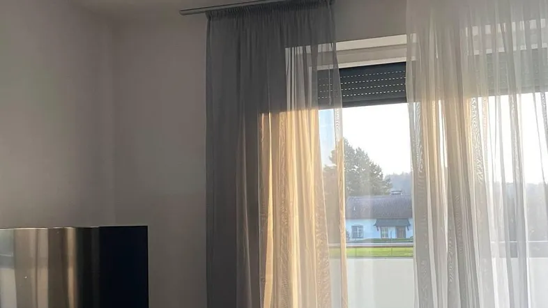 Helles Wohnzimmer mit Fernseher, Sofa und Zugang zu einem Balkon, durch den Sonnenlicht fällt.