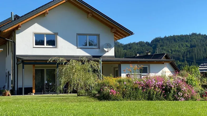 Hausfassade mit blühenden Blumenbeeten und weitem Blick auf die umliegende Natur.