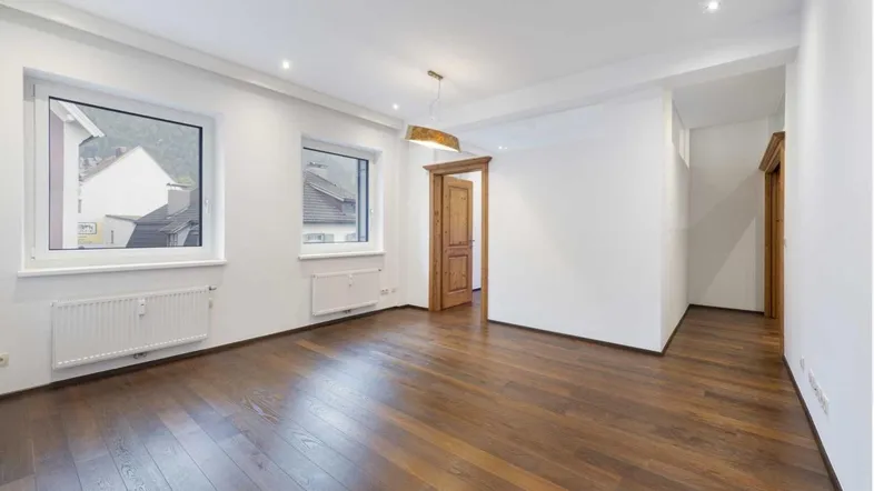 Helles, leeres Zimmer mit dunklem Holzboden und zwei Fenstern, bereit zur individuellen Gestaltung.