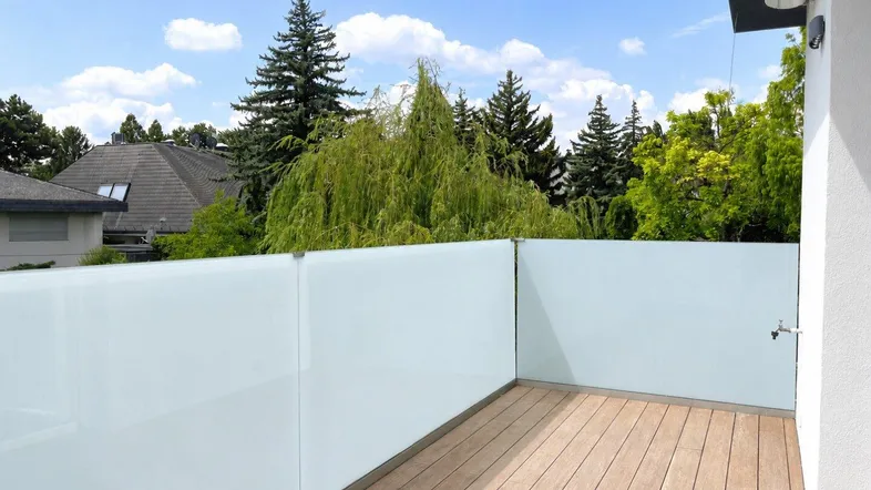 Heller Balkon mit Holzboden und moderner Glasbrüstung, umgeben von üppigem Grün.
