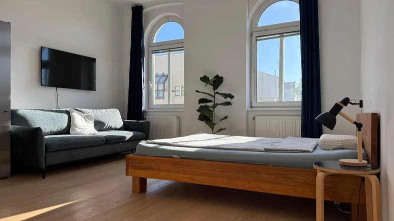 Helles Studio-Apartment mit Holzbett, Sofa und großen Rundbogenfenstern, die viel Tageslicht hereinlassen.