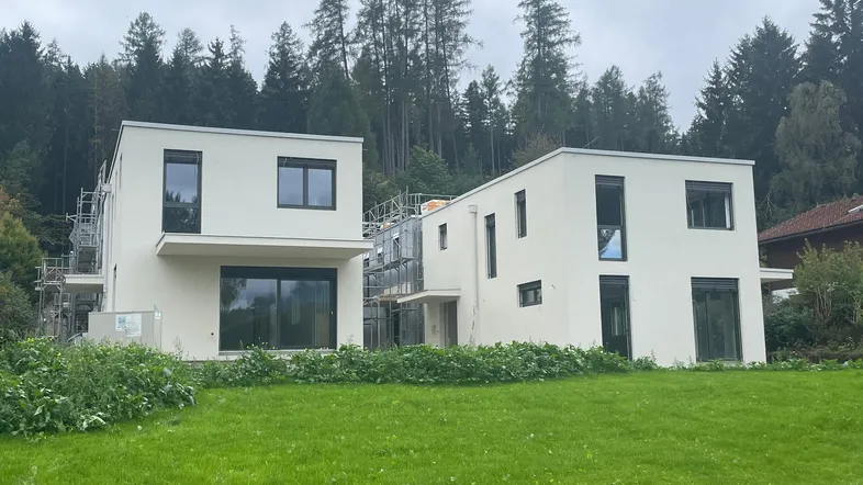 Zwei moderne Häuser im Bau mit weißer Fassade und großen Fenstern, umgeben von Grünflächen.