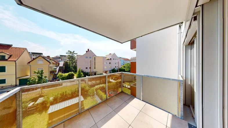 Heller Balkon mit Fliesenboden und Glasgeländer, bietet einen Ausblick auf die umliegenden Häuser und Gärten.